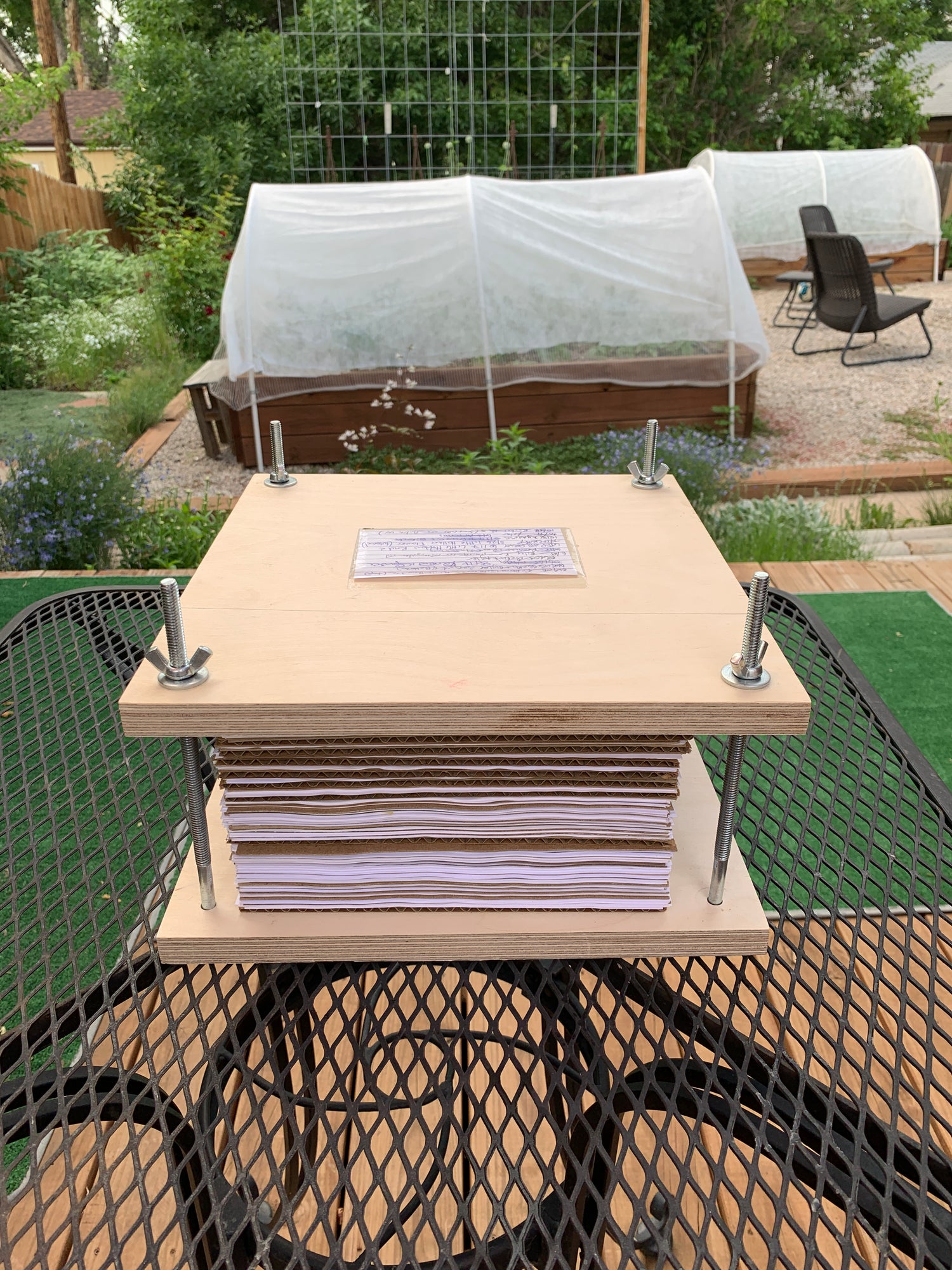 A wood flower press with paper and cardboard inside of it sitting on a iron table with garden beds in the background.