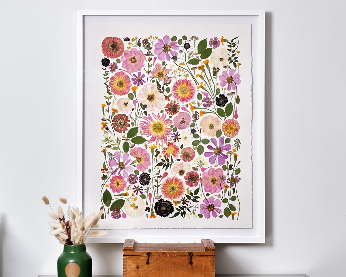 Colorful pressed flowers glued on white paper framed in a white frame that sits on a wood stand.