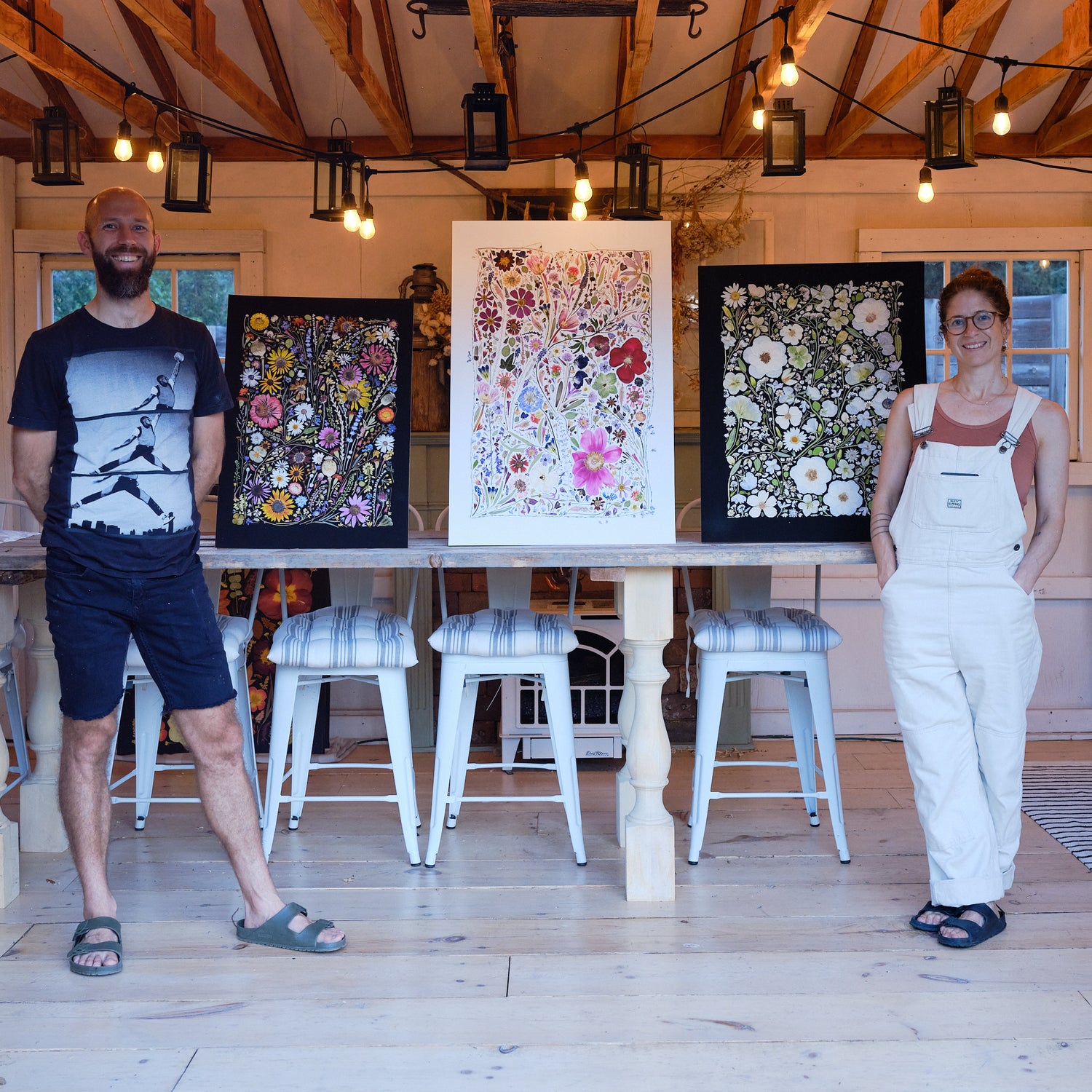A man and a woman standing in a barn with three pieces of pressed flower art.